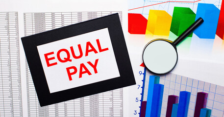 On the table there are reports of multi-colored charts, a magnifying glass and a sheet of paper in a black frame with the text EQUAL PAY. Business concept