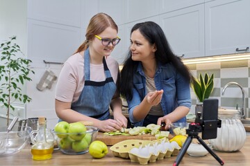 Mom and teen daughter cooking apple pie together, looking at smartphone screen