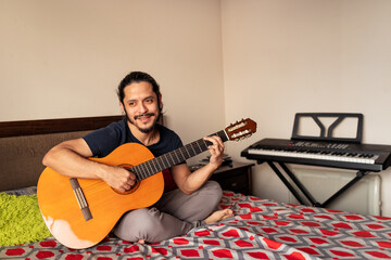 Young latin man playing acoustic guitar on bed in the bedroom at home