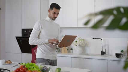 Busy Middle Eastern concentrated man walking with laptop in kitchen messaging online. Confident handsome focused guy typing on keyboard distracted from cooking of healthy salad indoors in home office