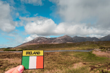 Tourist holding badge with sign Ireland and National flag of Ireland in focus. Mountains out of...