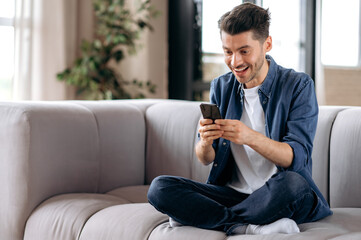 Obraz premium Excited young adult stylish caucasian guy, sitting at home in the living room on a couch, using his smartphone, texting with friends, reading good news, browsing the internet, winning lottery or game