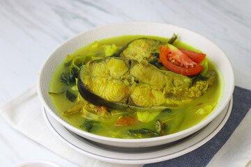 Pindang Ikan Patin or Catfish vegetables Soup , Indonesian food popular from Palembang. Served on ceramic bowl garnished with tomato slices. White marble background.