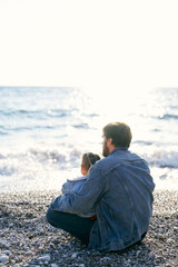 Dad in a denim jacket sits on a pebble beach and holds a little girl in his lap. Back view