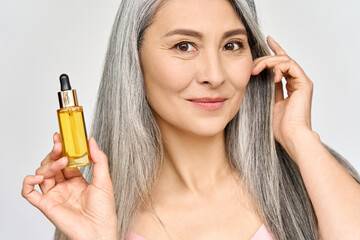 Happy smiling mid age Asian attractive woman with grey silky shiny volume hair holding bottle with serum essence rich nourishing oil for skincare and haircare. Cosmetics hair face treatment concept.