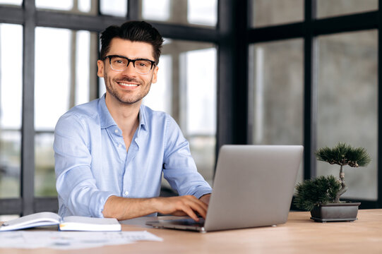 Portrait of a handsome intelligent caucasian man with glasses, businessman or manager, in formal shirt, sitting at a table with laptop, looking at camera and smiling friendly