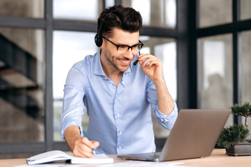 A friendly caucasian man with glasses, office worker or consultant, stands near a workplace, uses a headset and laptop for online consultation or negotiations, communicates via video communication