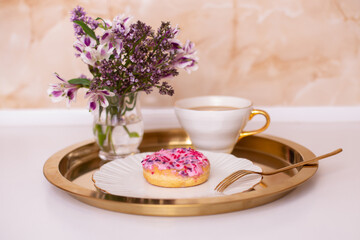 Coffee cup and sweet strawberry donut on golden plate with flower bouquet