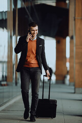 Going to airport terminal. Confident businessman traveler walking on city streets and pulling his suitcase drinking coffee and speaking on smartphone