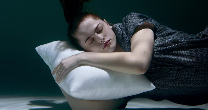 FALLING ASLEEP Young beautiful woman in pyjamas sinking deep under water with head on white pillow dreaming slow motion.