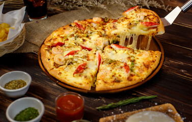 Selective focus and close up on a piece of double cheese homemade seafood pizza on spatula or scoop with blur background of wooden table, sackcloth, ingredients and fried potatoes. Food Concept.