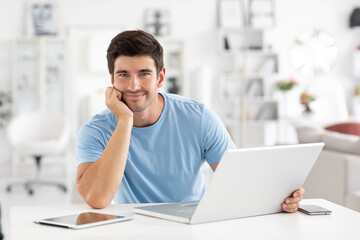 Handsome cheerful young male copywriter uses modern laptop for work, wears a blue t-shirt, look at...