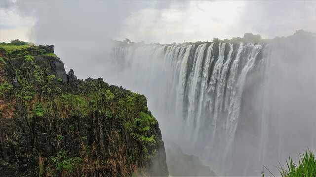 A Thick Fog From The Collapsing Streams Of Water Stands Over The Gorge. In The Foreground Is A Wet Rock With Green Vegetation. Tiny Figures Of People Are Visible On The Plateau. Victoria Falls. Zambia