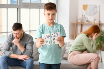 Sad little boy with torn drawing of family after quarrel between his parents at home