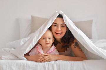 Happy woman and her little daughter under blanket on bed