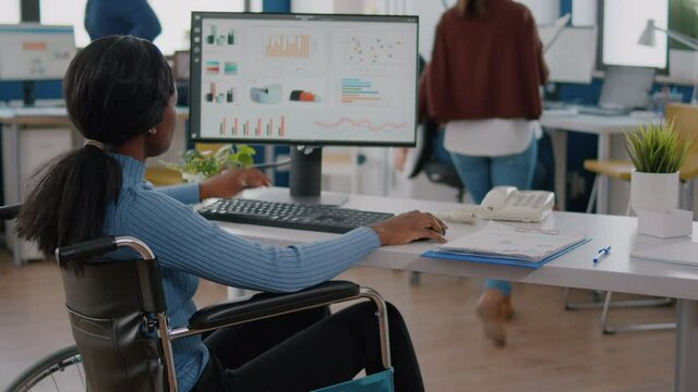 Busy focused african invalid paralized woman manager analysing graphics and financial statistics from computer, typing working in start-up business office sitting in wheelchair using modern technology