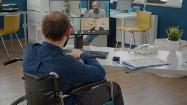 Back view of handicapped paralysed invalid manager talking with team on videocall about sale report, having video conference working in start-up office. Team using online meeting discussing on webcam