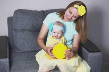 Little girl in yellow clothes and mom in yellow skirt are posing sitting in a gray chair playing with a yellow ball