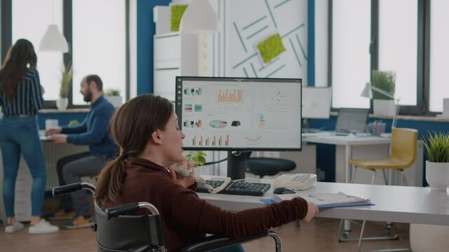 Disabled invalid paralysed businesswoman discussing business startegy with coworker at phone working from business office sitting in wheelchair. Handicapped employee explaning financial statistics