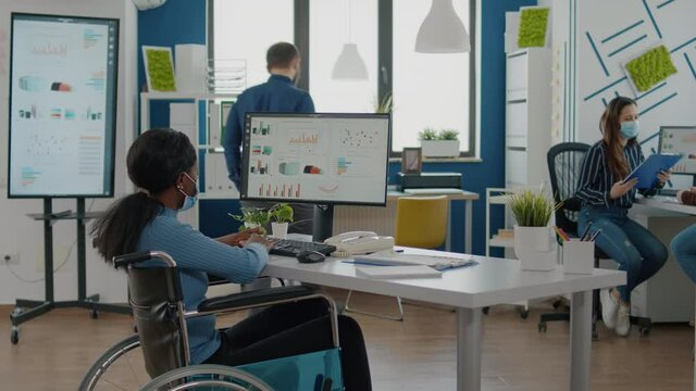 Paralysed black businesswoman and employee with protective mask working together at financial project in new normal start up company, compering statistics from documents looking at computer