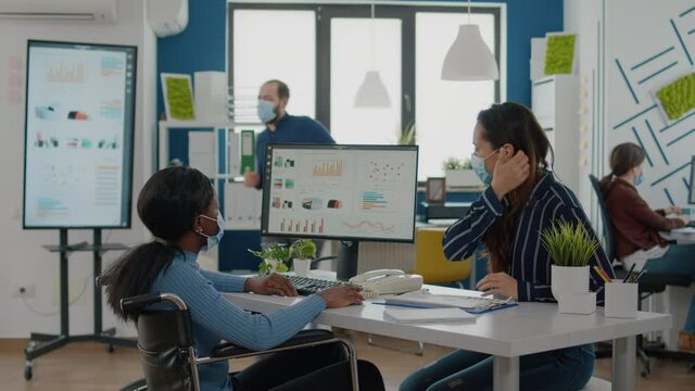 Invalid african businesswoman and employee with protective masks working together at financial project for start up company, compering statistics looking at computer discussing searching solution