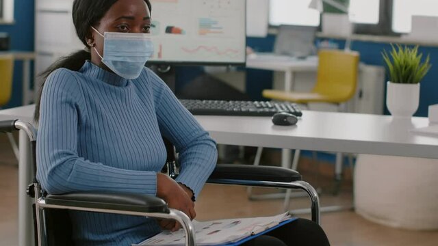African invalid handicapped woman employee with protection mask sitting immobilized paralized in wheelchair looking lost, new normal business office during covid 19 global pandemic working with team