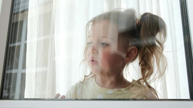 Pretty little girl waves hand looking out of window at home