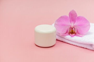 Fototapeta na wymiar Jar of moisturizing body cream, bath towel, orchid flower on a pastel pink background. Skin care cosmetic concept. Soft selective focus. Copy space.