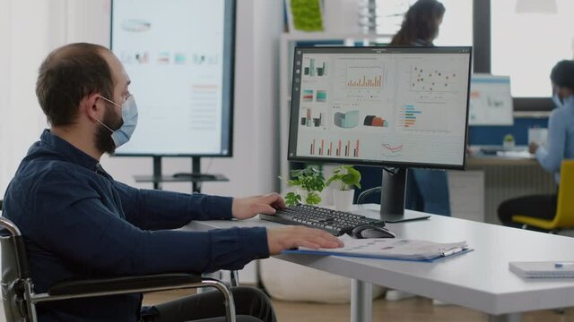 Immobilized business man with protective mask working in new normal business financial company typing on pc, checking reports, analysing data looking at desktop sitting in wheelchair