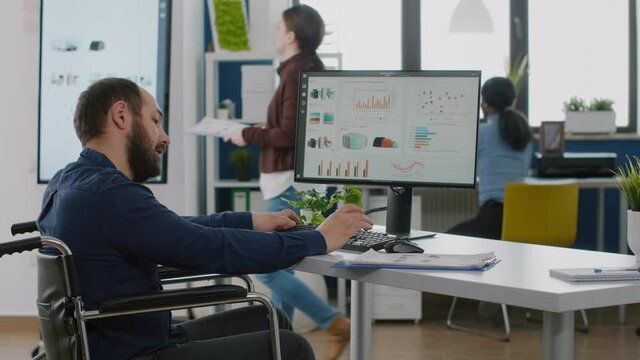 Disabled immobilized invalid businessman working at financial project respecting deadline typing on computer analysing statistic reports from documents sitting in wheelchair in start up office