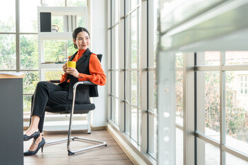Fototapeta na wymiar Portrait of Asian female manager or employee sitting at working desk with drink of coffee. smiling and cheerful woman in the office. copy space
