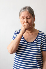 An elderly woman, a gray-haired lady of the 60s in a striped white and blue dress, does not feel well and coughs as a symptom of a cold or bronchitis. Healthcare concept.