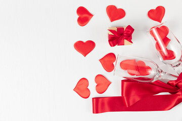 two champagne glasses, a box with a gift, a red silk ribbon and a lot of hearts scattered on a white wooden table. close-up