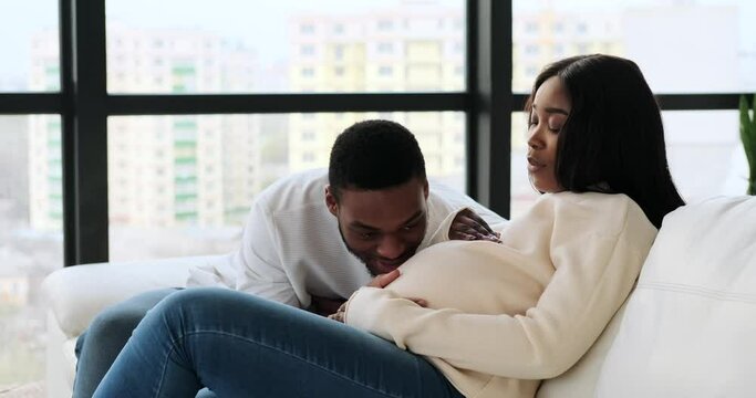 Man caressing and kissing belly of his pregnant wife sitting on sofa at home