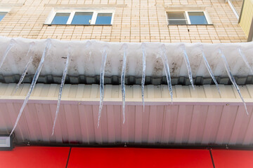 Long icicles hang dangerously down from the roof. The wall of the building is finished with red and white plastic tiles. Abnormal weather concept. New Year and Christmas is over.