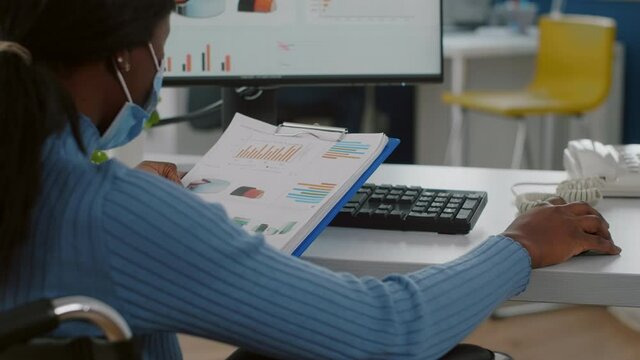 Close up of black paralysed woman worker with protective mask immobilized in wheelchair working on computer in new normal start up business office checking reports, analysing datas from clipboard