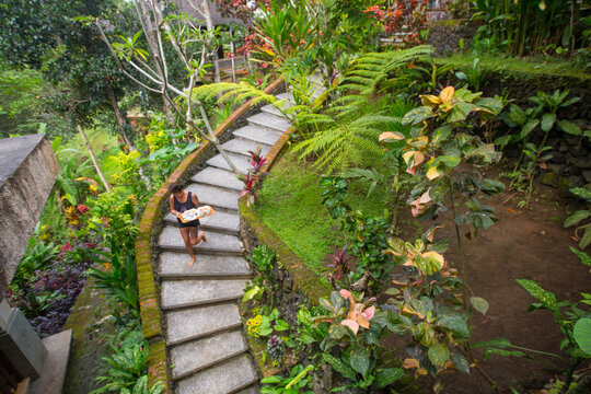 Woman carrying a breakfast tray of fruit and coffee down curved stair case through a lush jungle to a bungalow in Bali.