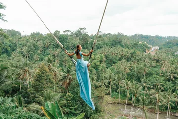 Fotobehang Tanned beautiful woman in a long turquoise dress with a train, riding on a swing. In the background, a rainforest and palm trees. Copy space. Rear view © _KUBE_