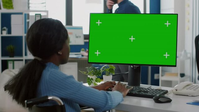Black paralyzed invalid businesswoman sitting in wheelchair using pc with green screen chroma key display, typing on computer while team planning project of strat up business