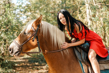 Fototapeta premium A brunette woman in a red dress sits astride a brown horse and smiles sweetly. Summer. Bright colors