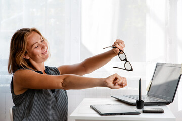 A woman sitting at a work Desk is warming up from fatigue. White window in the background. The...