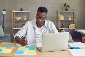 Young african american man studying or working on laptop computer from home sitting at desk writing...