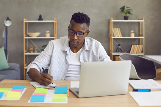 Young African American Man Studying Or Working On Laptop Computer From Home Sitting At Desk Writing Notes In Notebook With Sticker. Busy Black Entrepreneur Developing Startup Project Doing Research