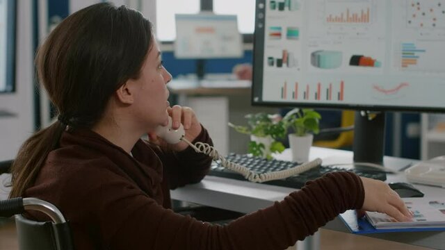 Paralysed financial woman expert sitting immobilized in wheelchair speaking at phone, working at pc in strat up company. Disabled invalid employee discussing with client of project, business partner