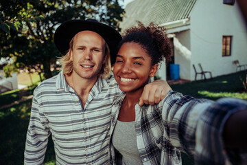Obraz premium Mixed race couple smiling taking selfie with cellular device standing in front of white farm home