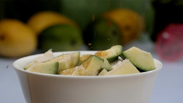 A sprinkling of spicy Masala on raw mangoes kept in a ceramic bowl - Aam Ka Achar. Yellow and green mangoes  with a small cocktail umbrella against a blurred background - Indian traditional pickle