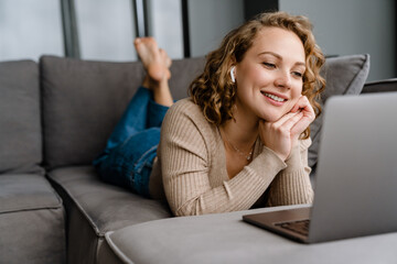 Young woman in earphones using laptop while lying on couch at home