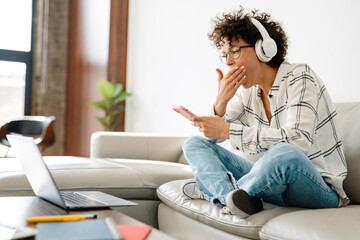 Young curly woman in headphones using laptop and cellphone at home
