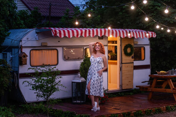 Young red-haired woman in white, floral sundress is resting by the motor at home in the evening. Trailer Track is decorated with garlands of lamps, blooming petunias. Happy girl smiles enjoys the trip