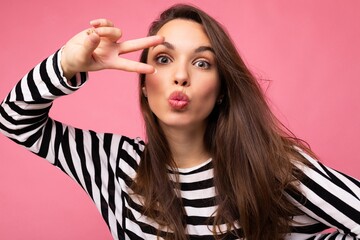 Obraz premium Photo of young european positive winsome sexy beautiful brunette woman with sincere emotions wearing casual striped pullover isolated on pink background with copy space and giving kiss
